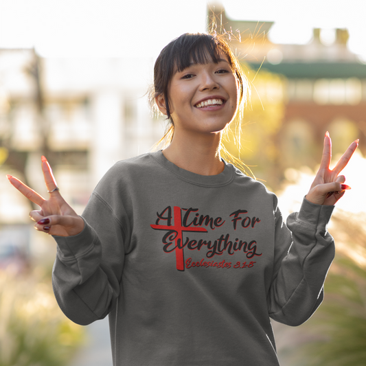 Young woman making peace signs while smiling outdoors and wearing a gray sweatshirt with a scripture design in red and black text with a Red Cross.